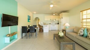 A modern living room with a sofa, coffee table, and TV. There is a dining area with chairs and a table. The kitchen is visible in the background with white cabinets and appliances. - Just off the Beach, Relax at Inn At Grace Bay #121 (Grace Bay)