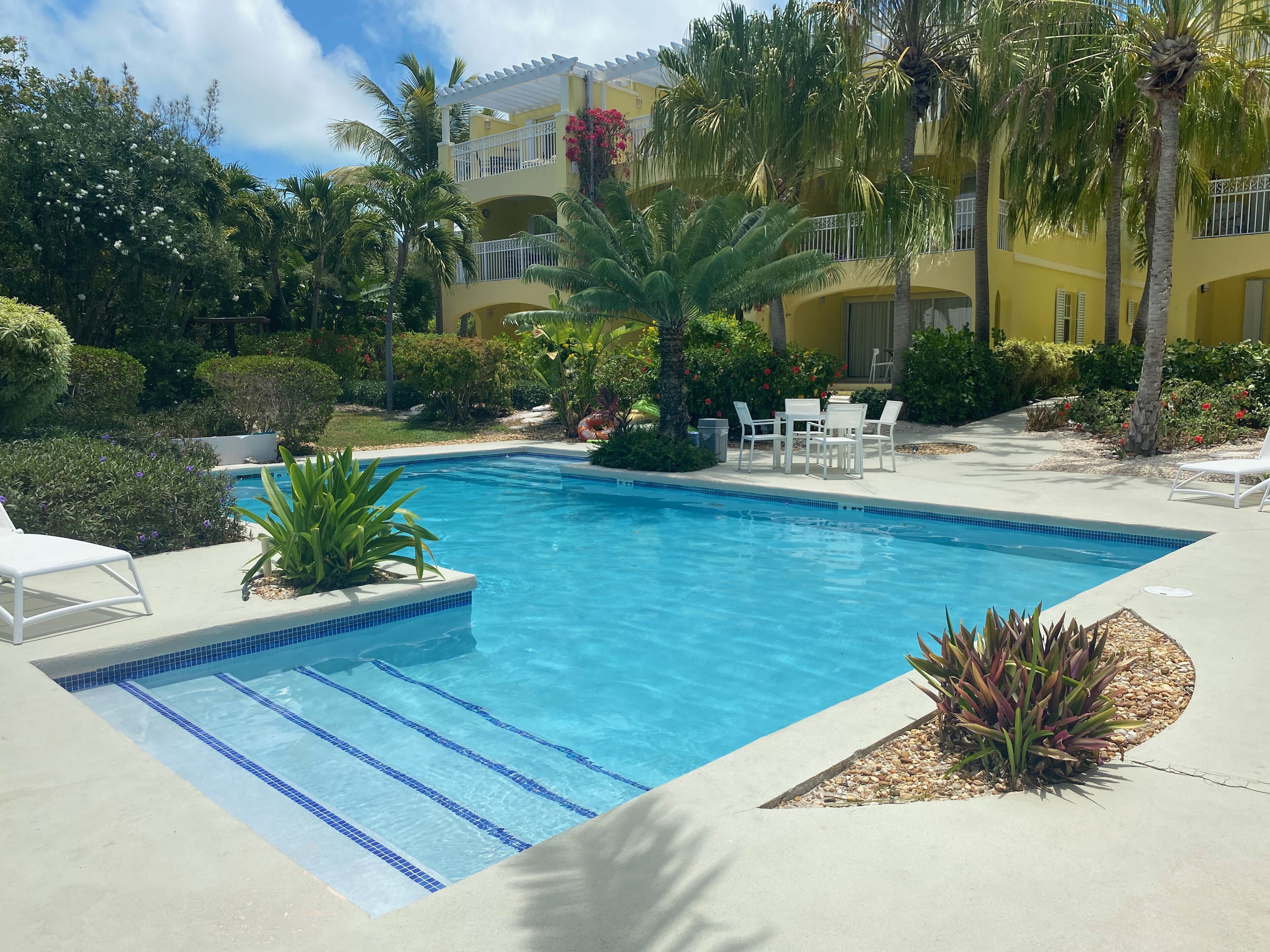 Une piscine à l’eau d’un bleu cristallin, entourée d’une terrasse en béton. On y trouve des chaises longues et une petite table avec des chaises. L’espace piscine est adjacent à un bâtiment doté de balcons et d’un jardin où poussent diverses plantes et fleurs.
