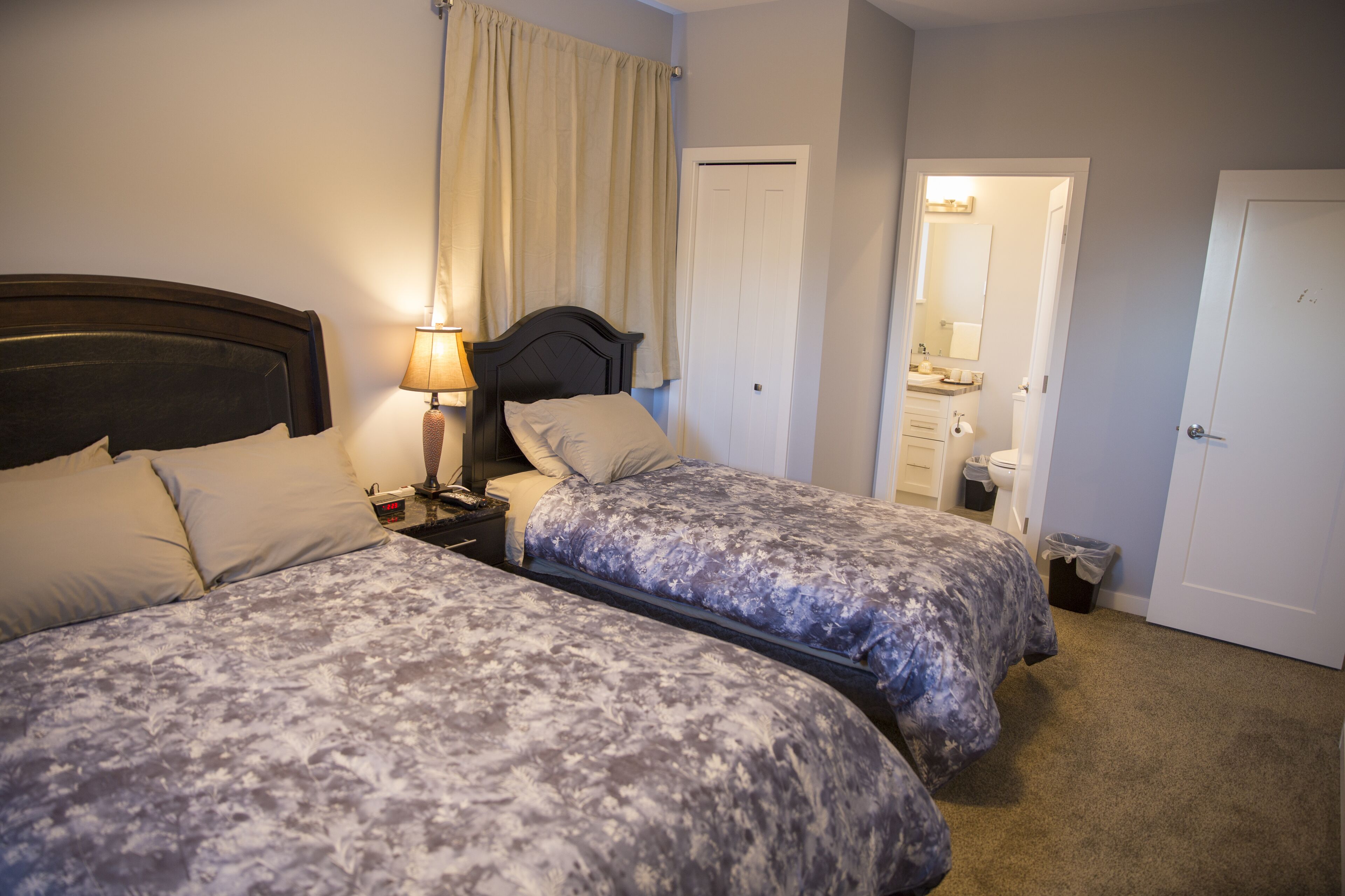 Une chambre d’hôtel avec deux lits, une table de chevet avec une lampe, une salle de bain visible à travers une porte ouverte et une poubelle.