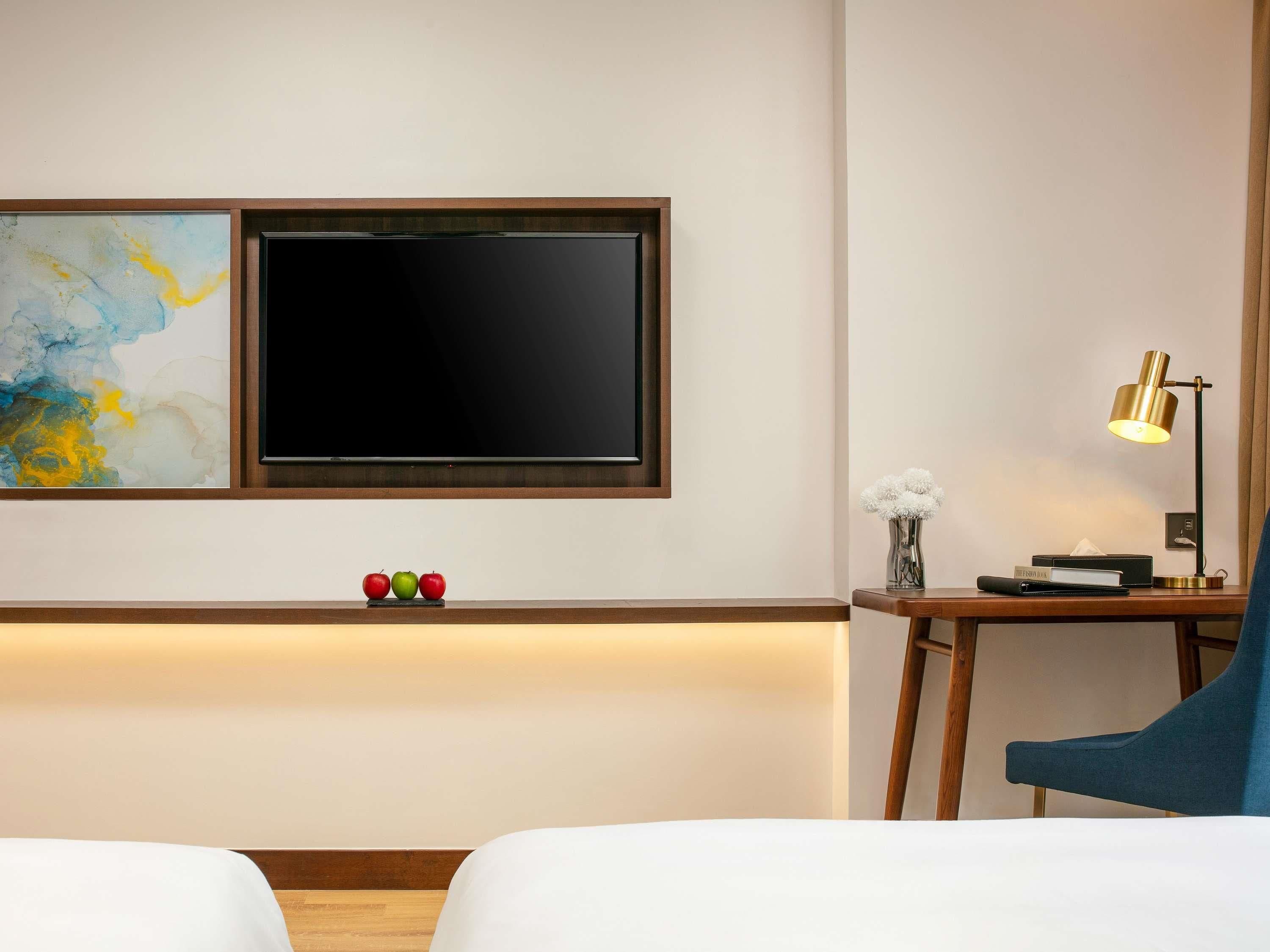 Habitación de hotel con una televisión de pantalla plana montada en la pared, una mesita de noche con lámpara, un jarrón con flores y una cama con ropa blanca.