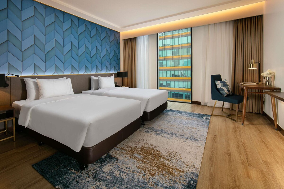 Een hotelkamer met twee bedden, een bureau, een stoel, een groot raam met uitzicht op een gebouw en een blauwe, bewerkte wand.
