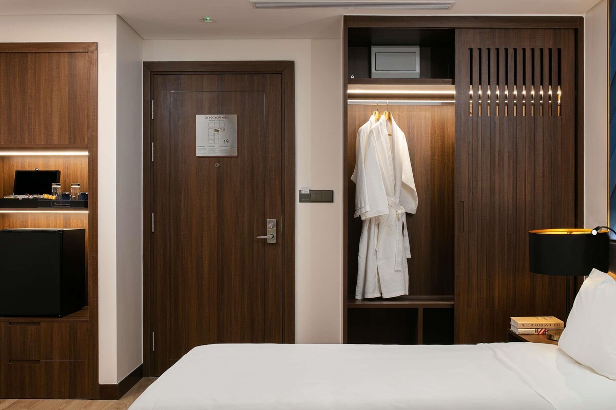 Een hotelkamer met een houten garderobe, een bed met witte lakens, een badjas hangend aan de garderobe en een nachtkastje met een lamp en een boek.