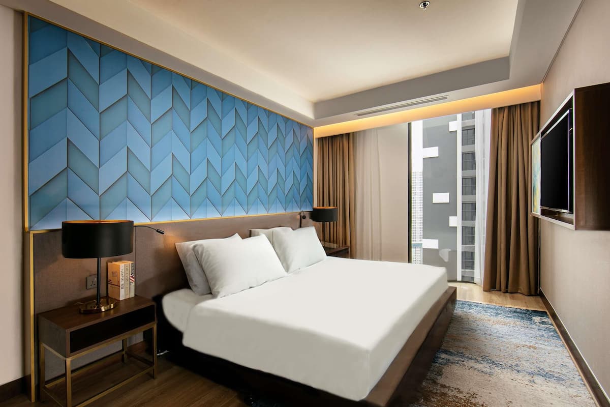 Habitación de hotel con una cama grande, una mesita de noche, una lámpara, un televisor y vistas a la ciudad.