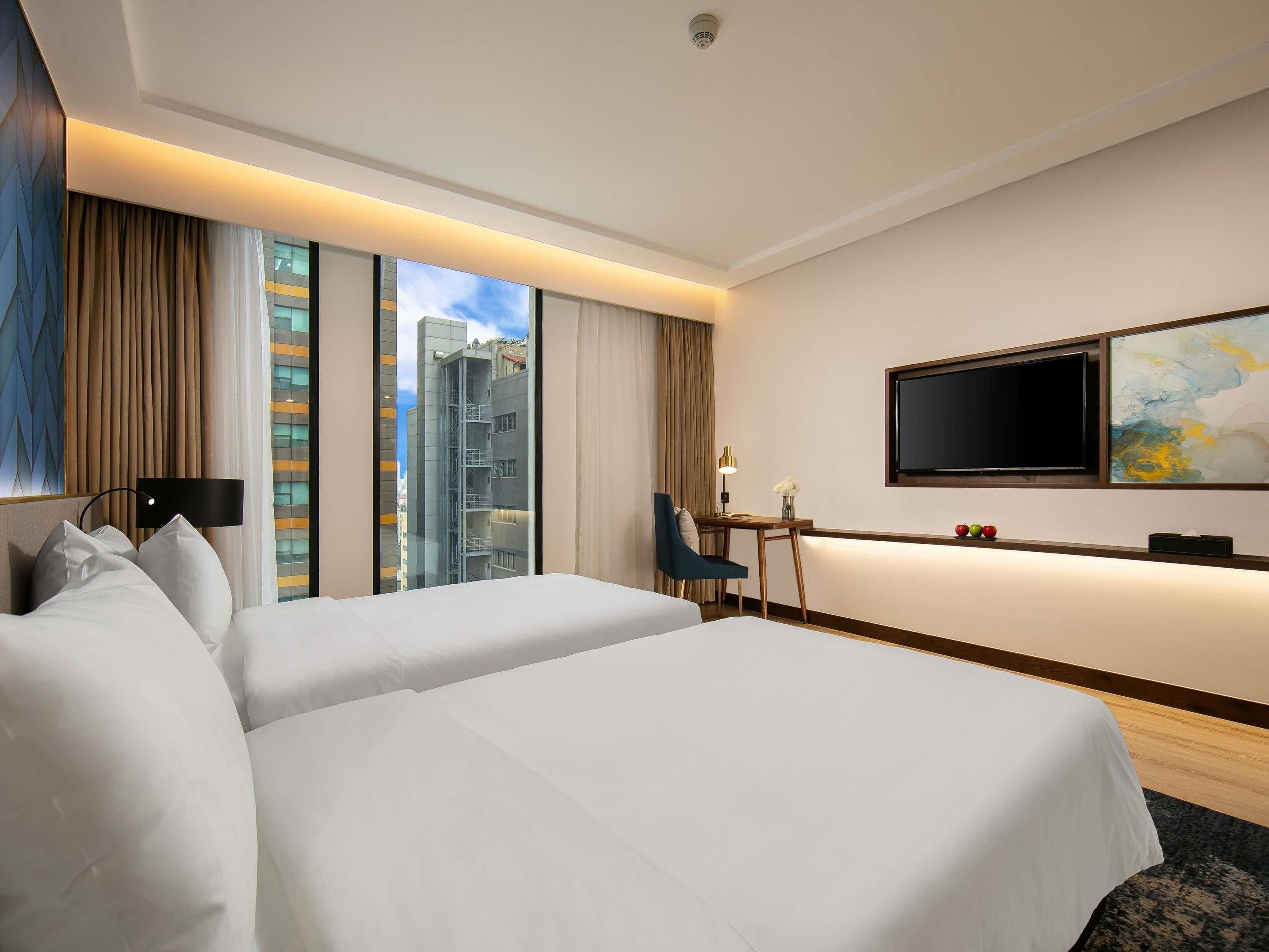 A hotel room with two beds, a desk, a chair, a TV, and a window with a city view.