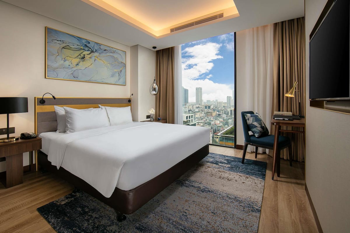 Habitación de hotel con una cama grande, un escritorio con silla, televisión y una vista a la ciudad, además de un cuadro en la pared.