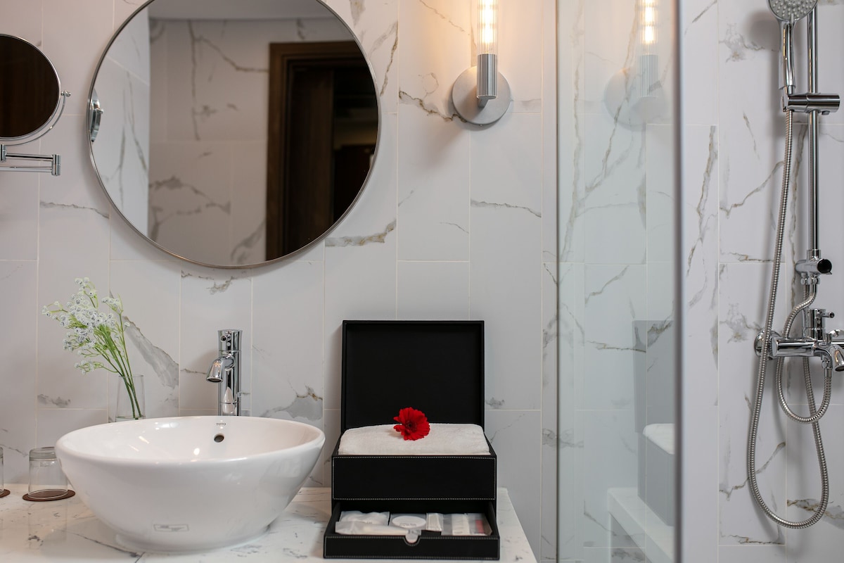 Une salle de bain avec un lavabo blanc, un miroir rond, une boîte noire ornée d’une fleur rouge et une douche.