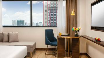 A hotel room with a bed, a desk, a chair, a window with a city view, and a TV.