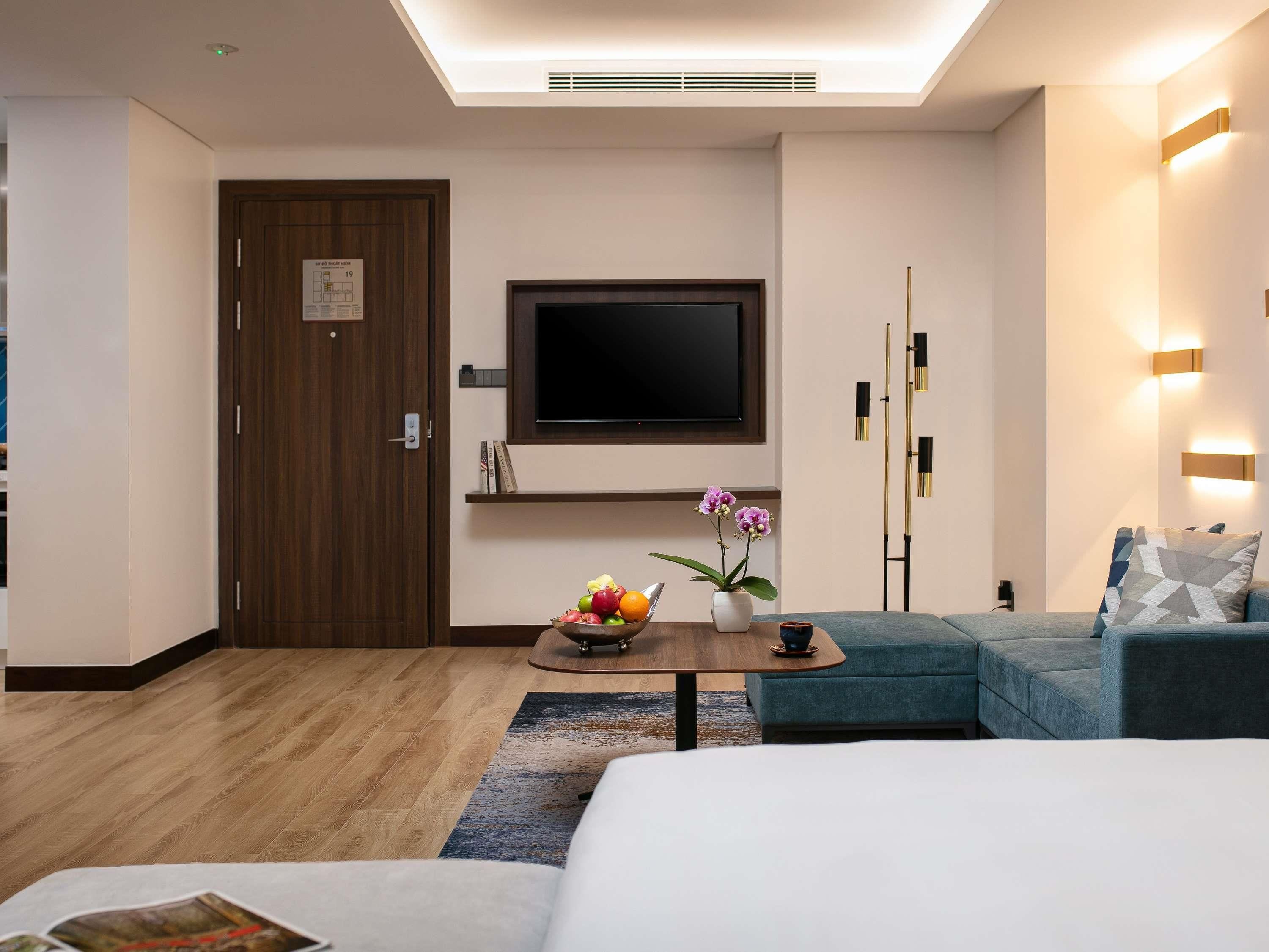 Una habitación de hotel moderna con televisor de pantalla plana, una puerta de madera, un sofá, una mesa de centro con fruta y flores, y una alfombra estampada.