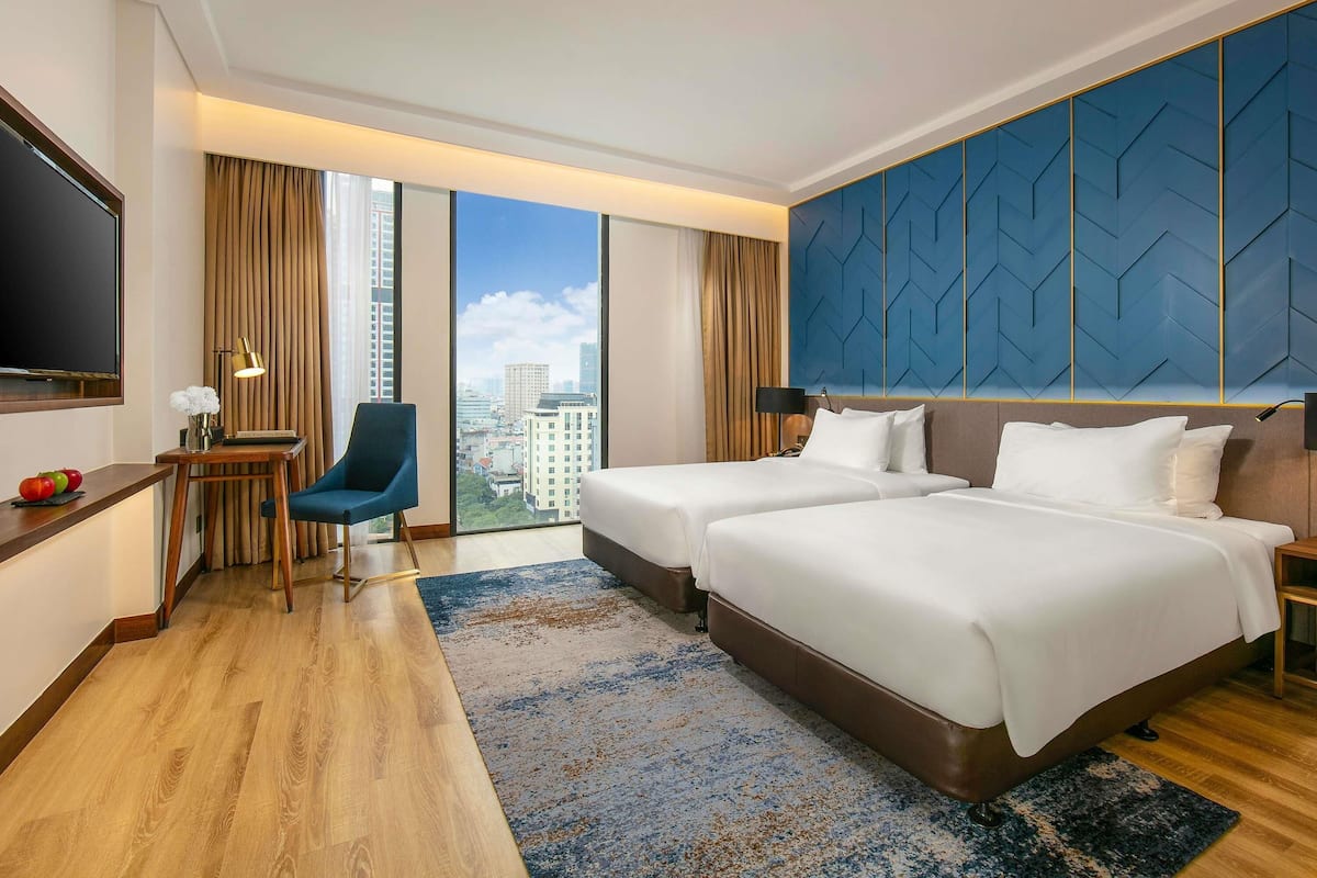 Habitación de hotel con dos camas, un escritorio con silla, televisión y un ventanal con vistas a la ciudad.