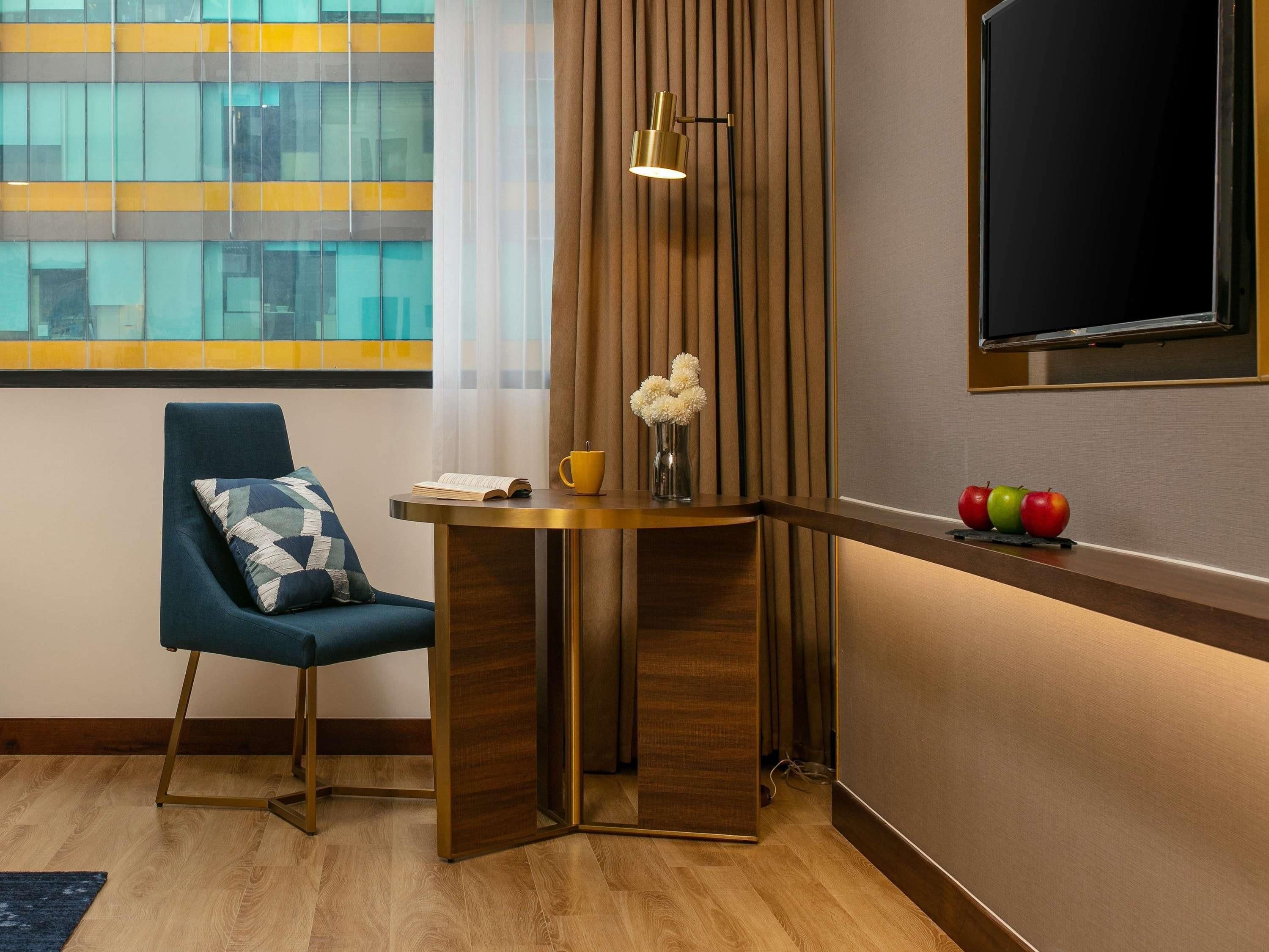 A modern hotel room with a blue armchair, a small round table with a book and a yellow mug, a vase with white flowers, and a TV mounted on the wall.