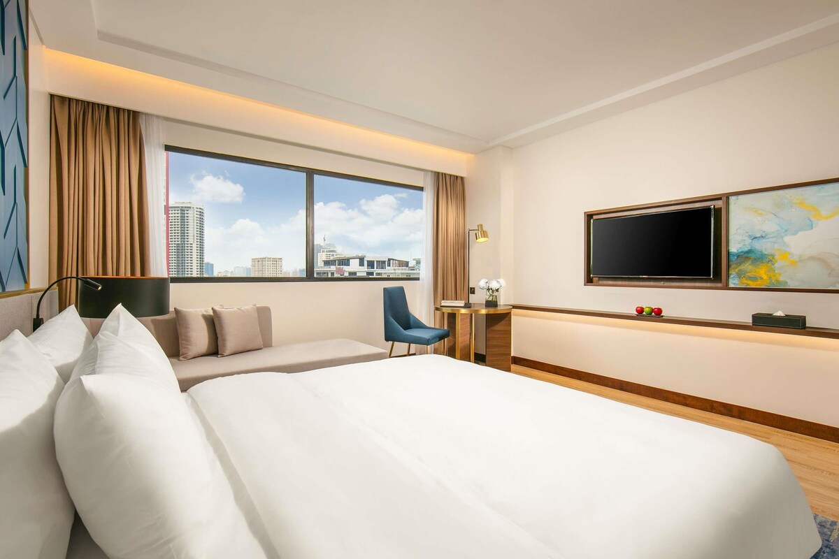 Habitación de hotel con una cama grande, un escritorio, una silla, un televisor y vistas a la ciudad.