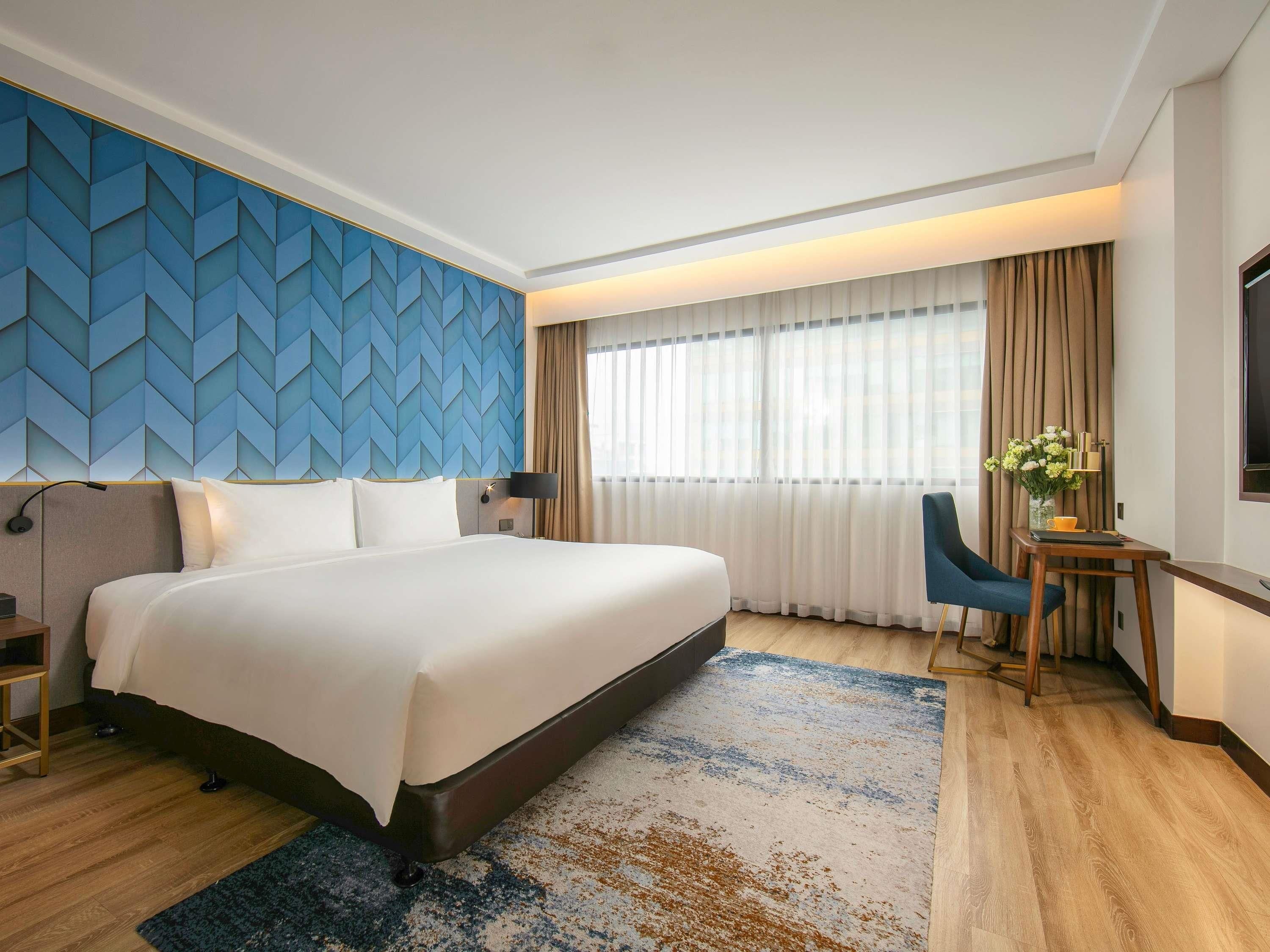 Quarto de hotel moderno com uma cama grande, uma escrivaninha, uma cadeira e vista para a cidade pela janela.