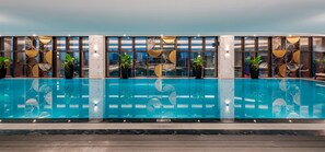 A modern indoor swimming pool with a city view, surrounded by potted plants and geometric wall designs. - Mövenpick Living West Hanoi (Hanoi)