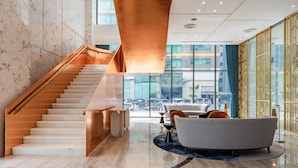 A modern hotel lobby with a marble staircase, a round coffee table, and a view of the city through large windows.