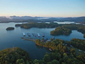 An aerial view of a lakeside property with several boats docked, surrounded by dense forest and hills in the background.