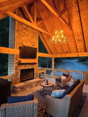Eine Holzterrasse mit Kamin aus Stein, Korbmöbeln und einem Fernseher.