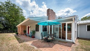 Une terrasse avec une table et des chaises, un parasol bleu et un chemin de briques menant à une maison dotée d’une cheminée.