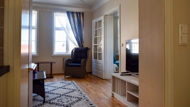 Una sala de estar con televisor, una silla de mimbre, un tapete azul y blanco, y una puerta que da a otra habitación.