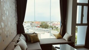 Uma sala de estar moderna com um sofá, almofadas e uma mesa de centro de vidro. Uma janela grande oferece vista para edifícios e árvores.