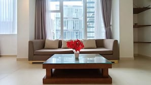 A modern living room with a beige sofa, a glass coffee table, and a view of tall buildings through large windows. - Glenwood Residences (Ho Chi Minh City)