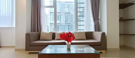 A modern living room with a beige sofa, a glass coffee table, and a view of tall buildings through large windows.