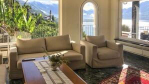 Uma sala de estar com sofá, poltrona e mesa de centro, vista para montanhas e água, e uma varanda com guarda-corpo.