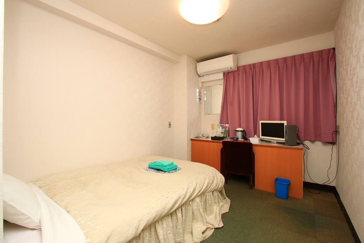 Hotellihuone, jossa on sänky, työpöytä tietokoneella, televisio ja ikkuna, jossa on verhot.