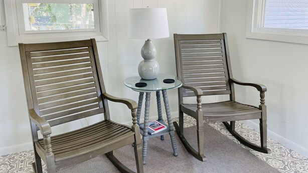 Deux fauteuils à bascule, une petite table en verre avec une lampe blanche et un tapis à motifs dans une pièce aux murs et aux fenêtres blancs.