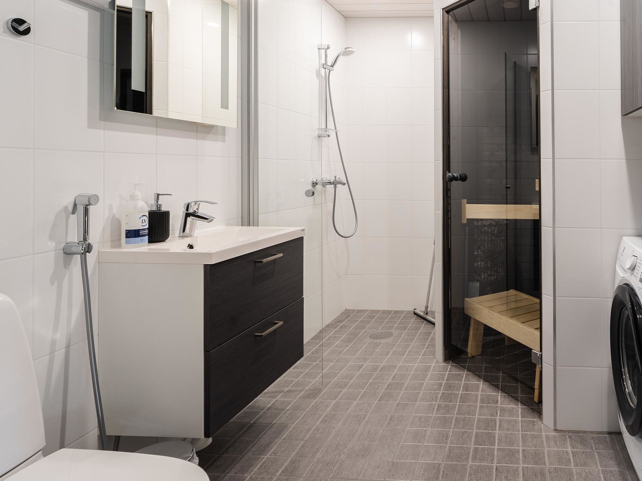 Un baño moderno con lavamanos blanco, mueble oscuro, ducha y un banco de madera.