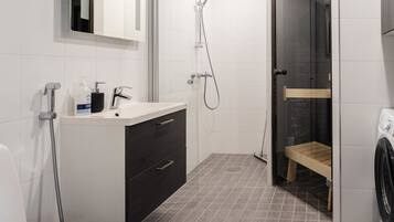 Un baño moderno con lavamanos blanco, mueble oscuro, ducha y un banco de madera.