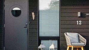 A black door with a circular window, a number '12' on the wall, a wicker chair, and lanterns on a wooden deck.