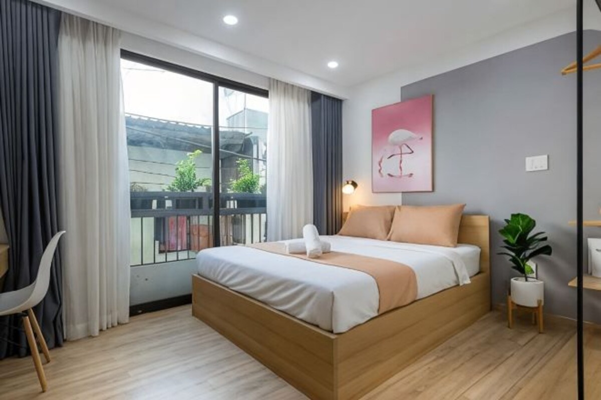 A modern hotel room with a large bed, a view of the outside, a small desk, and a potted plant.
