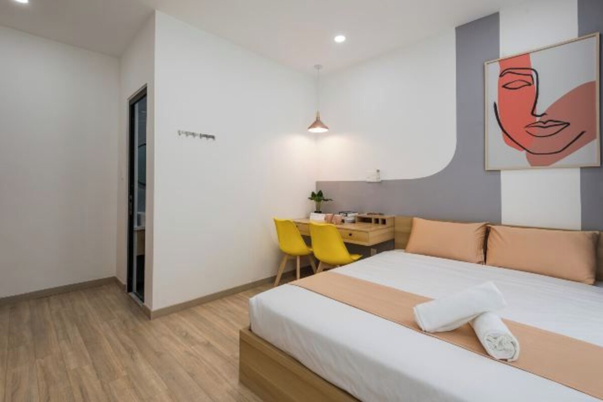 A modern hotel room with a bed, a desk, and a chair. There is a painting on the wall and a plant on the desk.