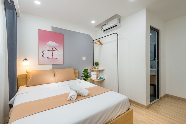 A neatly made bed with a beige blanket and a white pillow, a wall-mounted air conditioner, a small shelf with a plant and a phone, and a door leading to a bathroom. - Cozrum Homes Ambera House (Ho Chi Minh City)