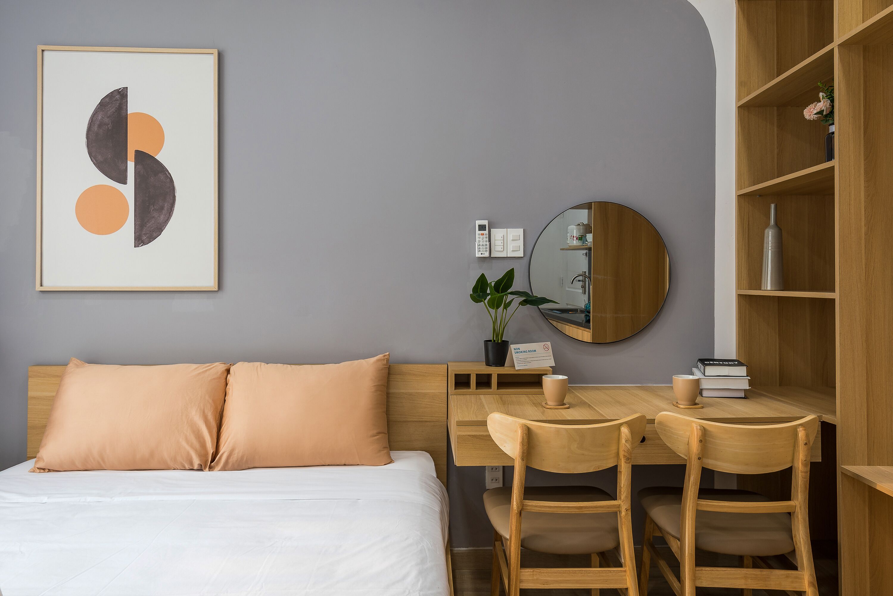 A neatly arranged hotel room with a bed, a desk with two chairs, a small table with a cup and books, a potted plant, and a framed artwork on the wall.