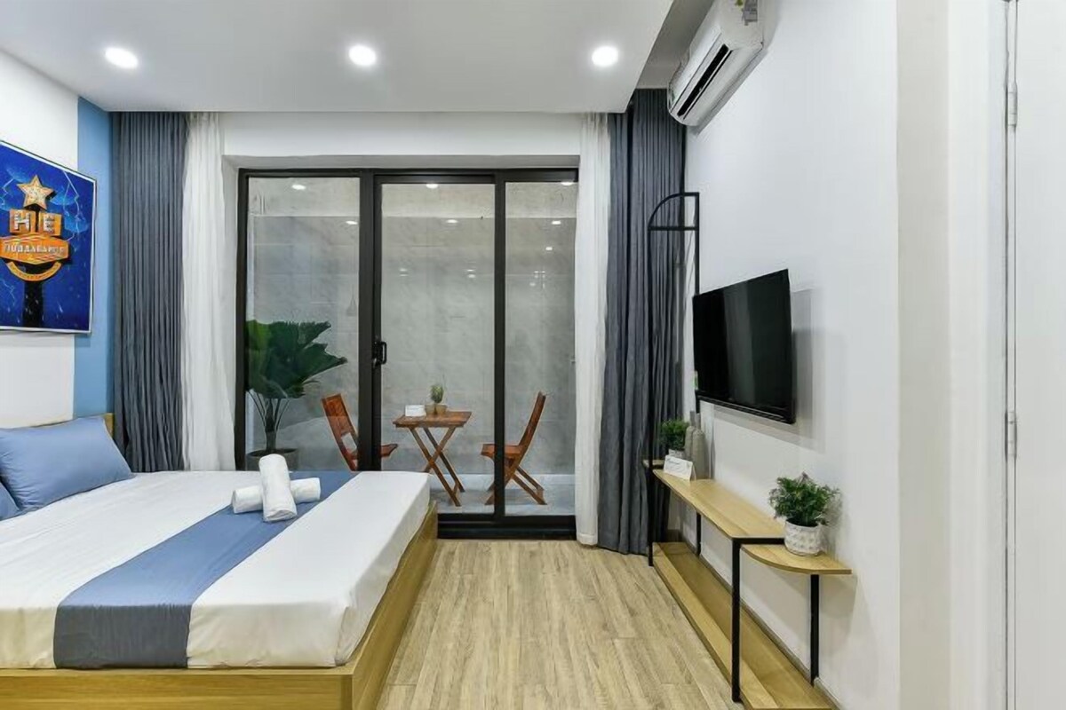 A hotel room with a bed, a TV, a small table with a plant, and a sliding glass door leading to a balcony.
