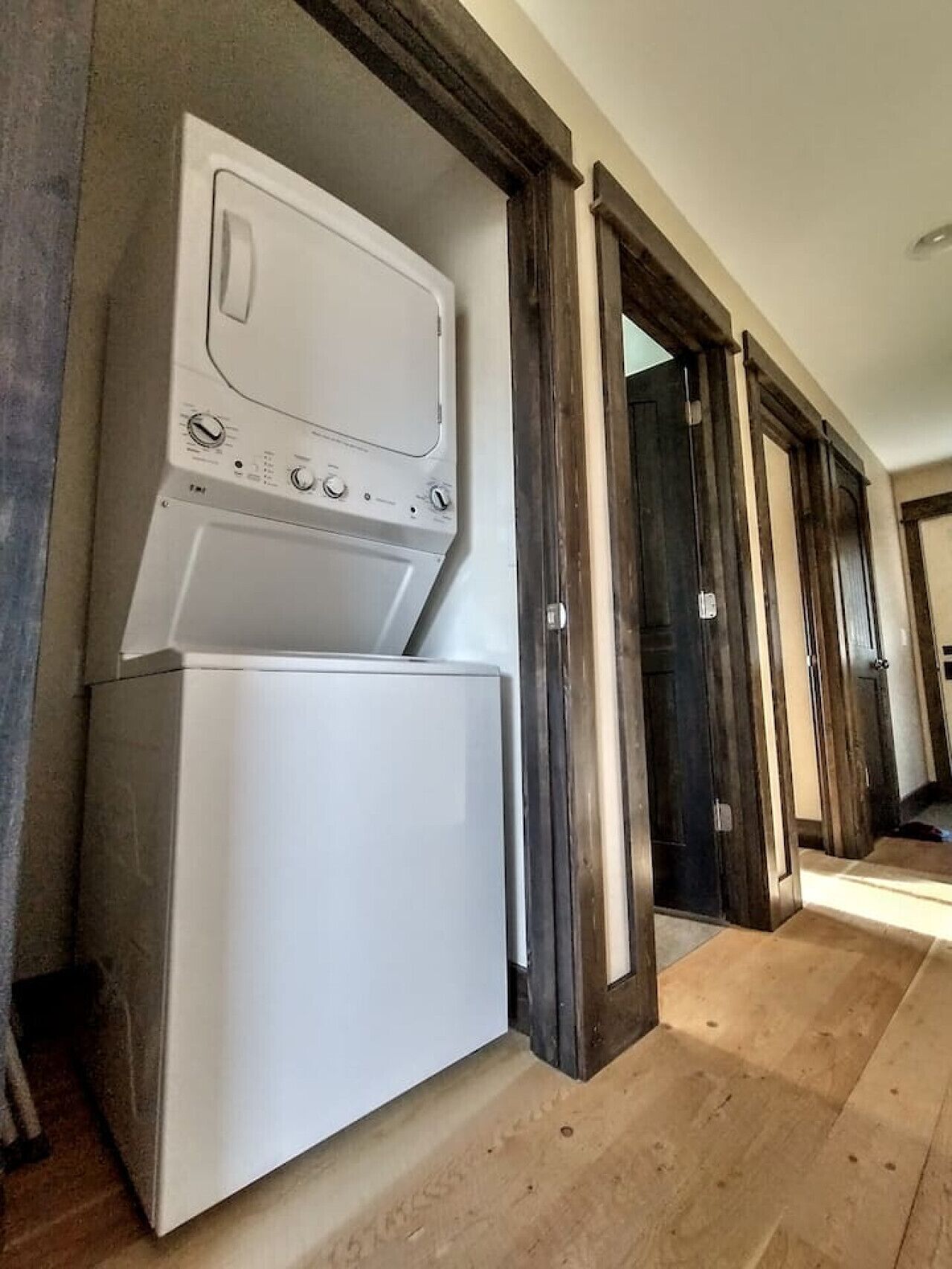 A laundry room with stacked appliances, a wooden floor, and dark wooden doors leading to other rooms.