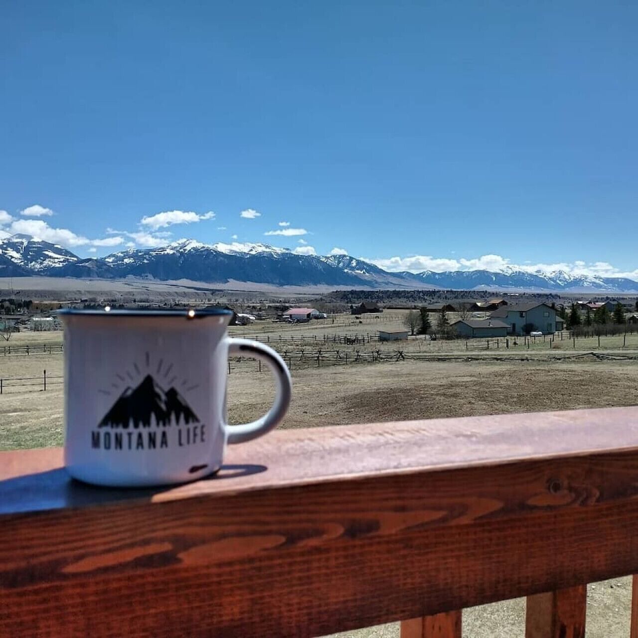 A white mug with a mountain design labeled "MONTANA LIFE" on a wooden railing with a view of snow-capped mountains and a rural landscape in the background.