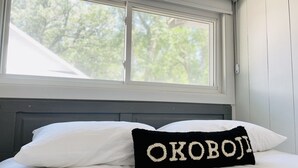 Una cama con almohadas blancas y una almohada negra con texto bordado, una ventana arriba y una pared blanca.