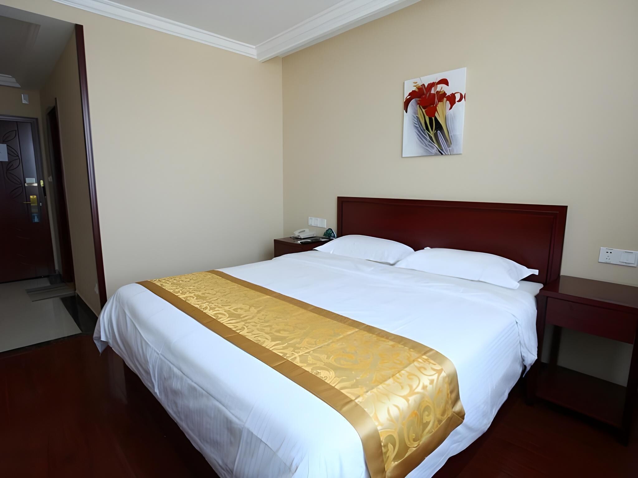 A hotel room with a large bed, a wooden headboard, a bedside table with a phone, and a painting of flowers on the wall.