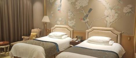 A hotel room with two beds, a chair, a lamp, and a wall with floral and bird artwork.