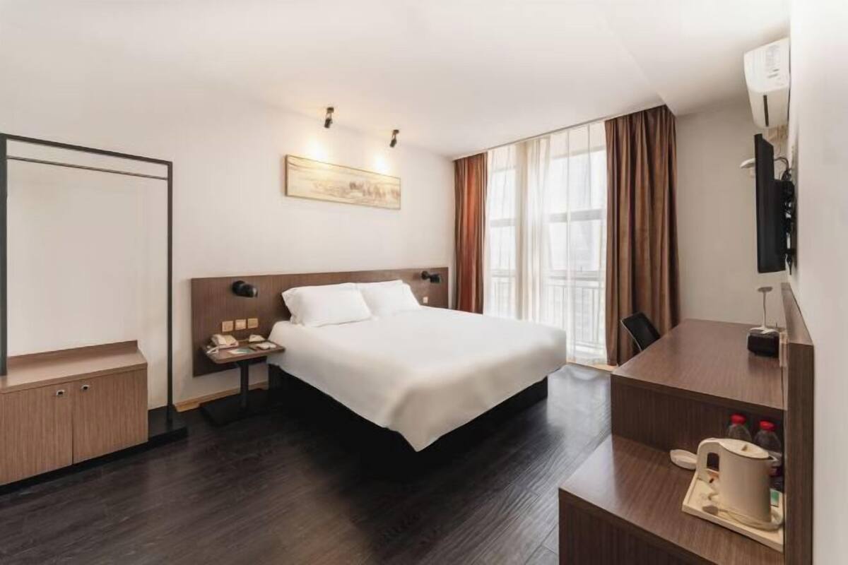 A hotel room with a large bed, a desk with a telephone and a coffee maker, a flat-screen TV mounted on the wall, and a window with curtains.