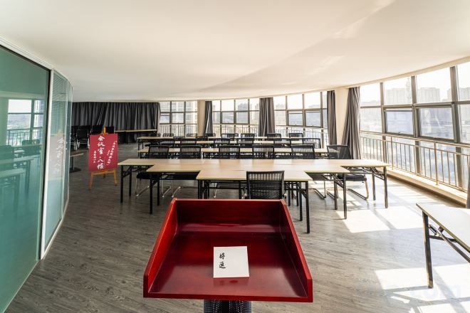 Una amplia sala de conferencias con varias mesas y sillas, ventanas grandes y una mesita roja.