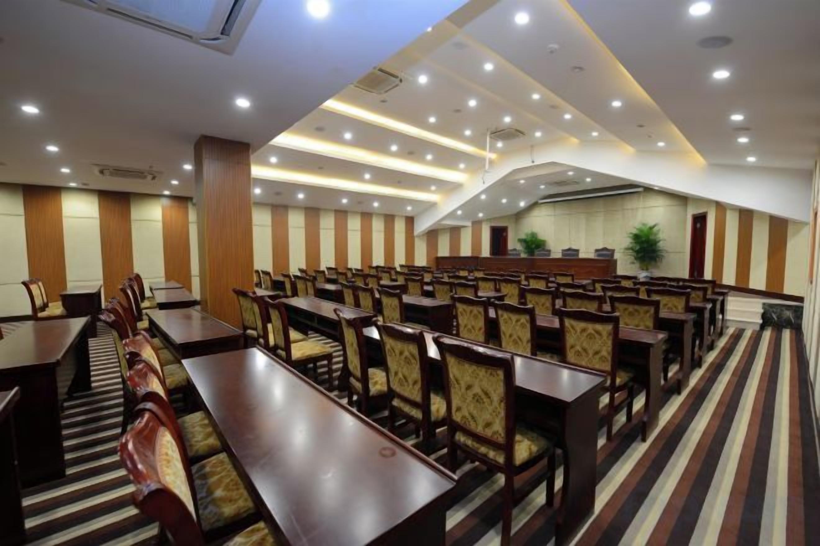 Une salle de conférence avec des rangées de tables et de chaises, un plafond haut doté d’un éclairage encastré et un tapis à rayures.