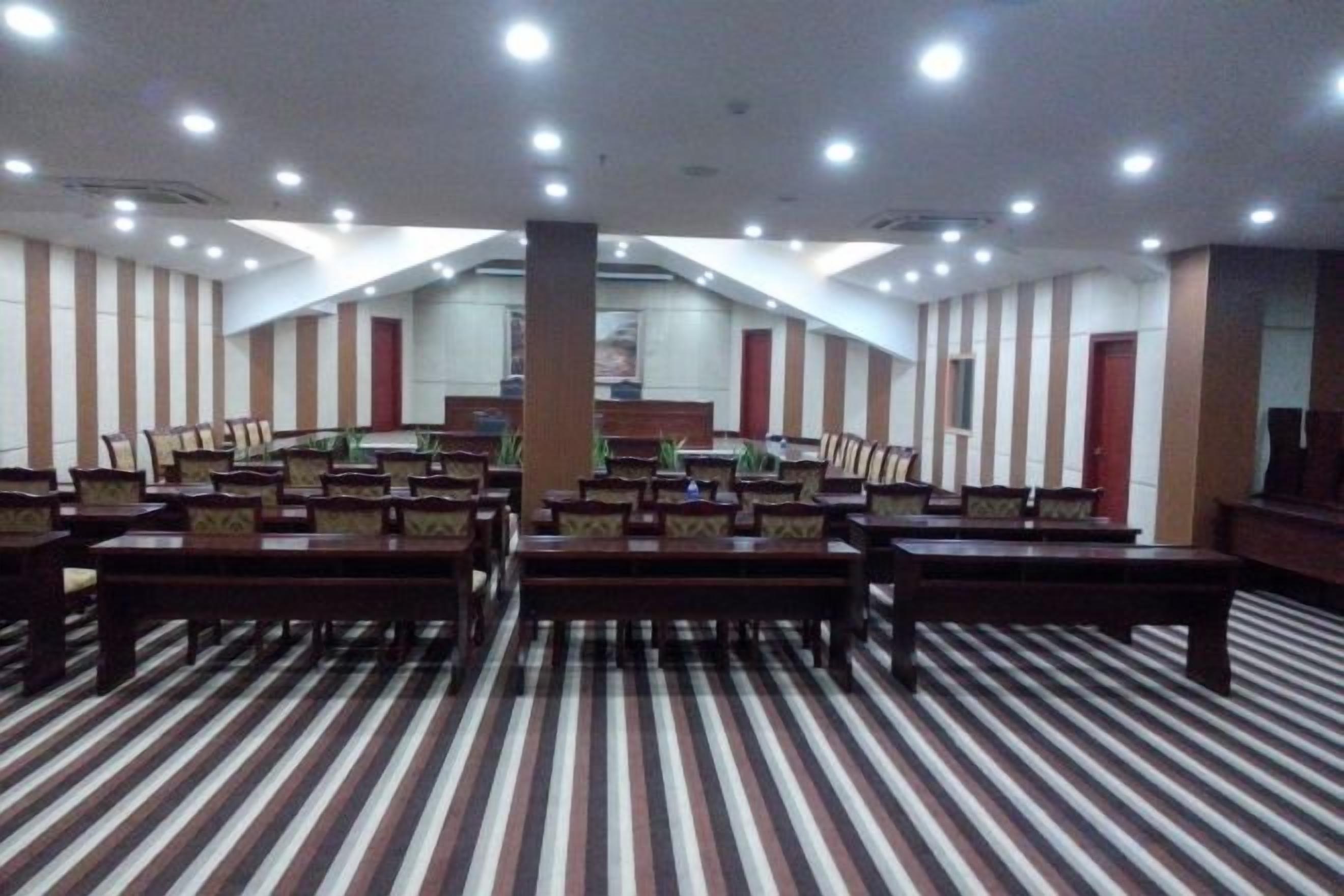 Sala de conferência com fileiras de cadeiras, uma mesa grande e teto alto com iluminação embutida.