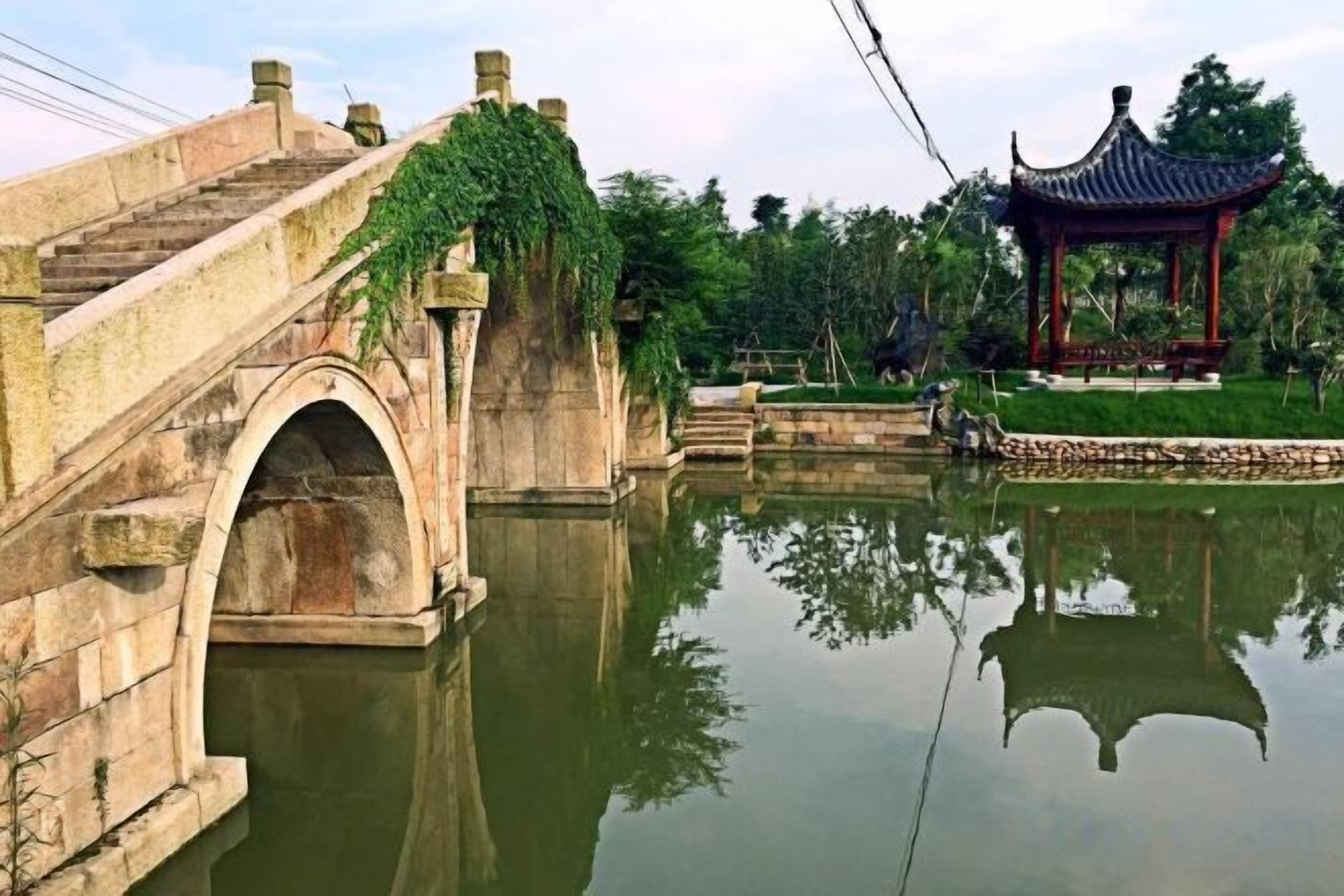 Un puente de piedra sobre aguas tranquilas, un pabellón tradicional y un reflejo nítido en el agua.