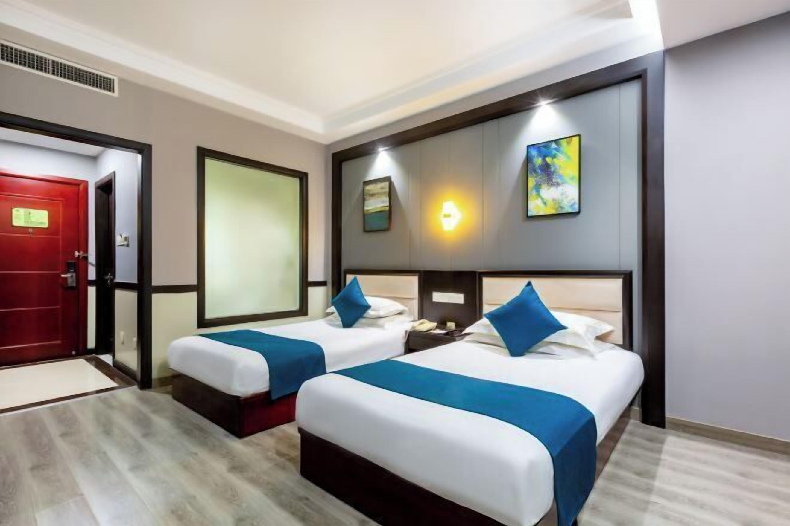 Une chambre d’hôtel avec deux lits, une porte rouge, un luminaire fixé au mur et un tableau.