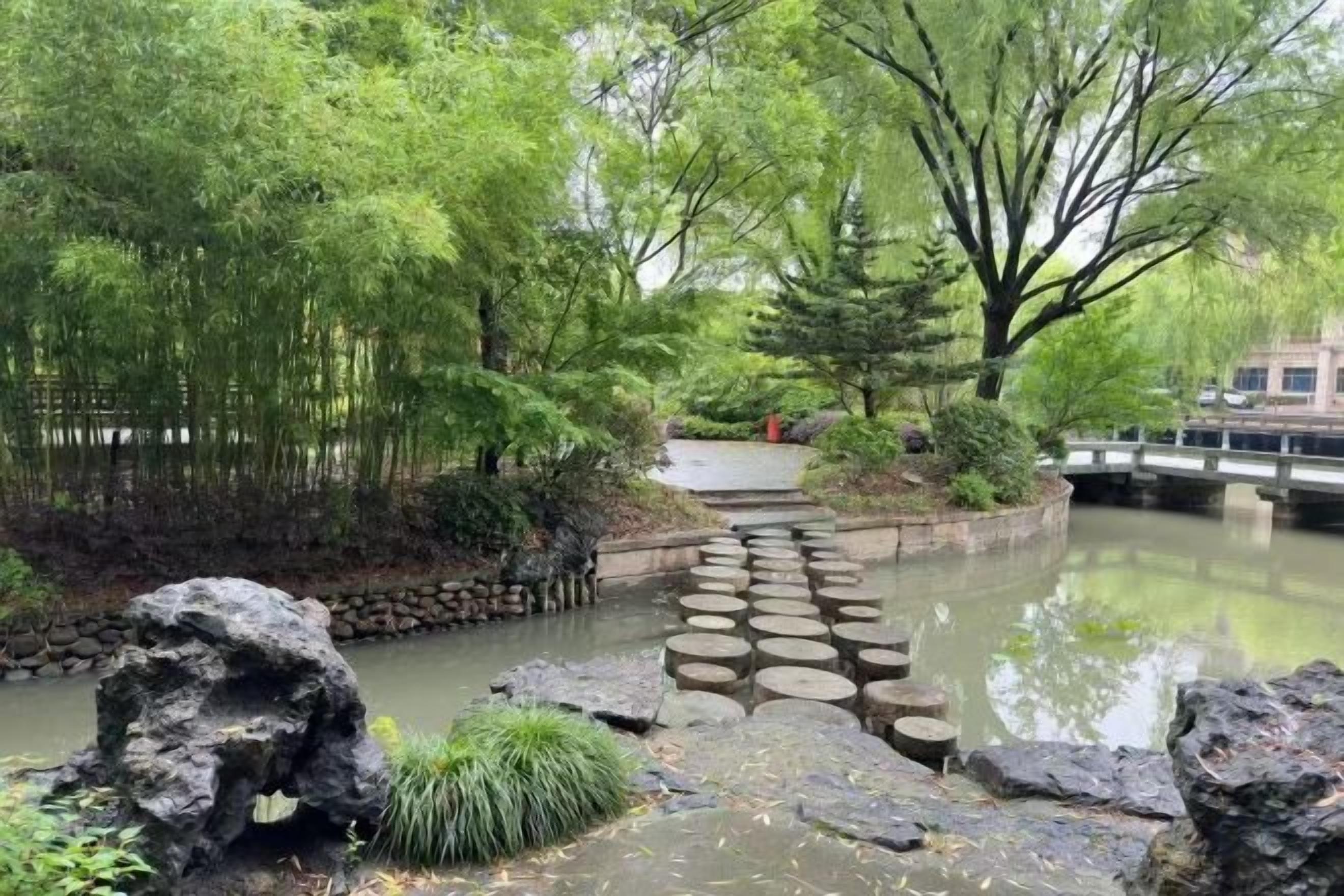 Un jardin paisible avec un pont de pierre enjambant un étang, le tout entouré d’une végétation luxuriante et d’arbres.