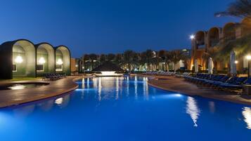 A nighttime swimming pool with lounge chairs and a building with arched windows in the background.