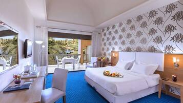 A hotel room with a large bed, a dining area with chairs and a table, a TV, and a view of a pool and palm trees.